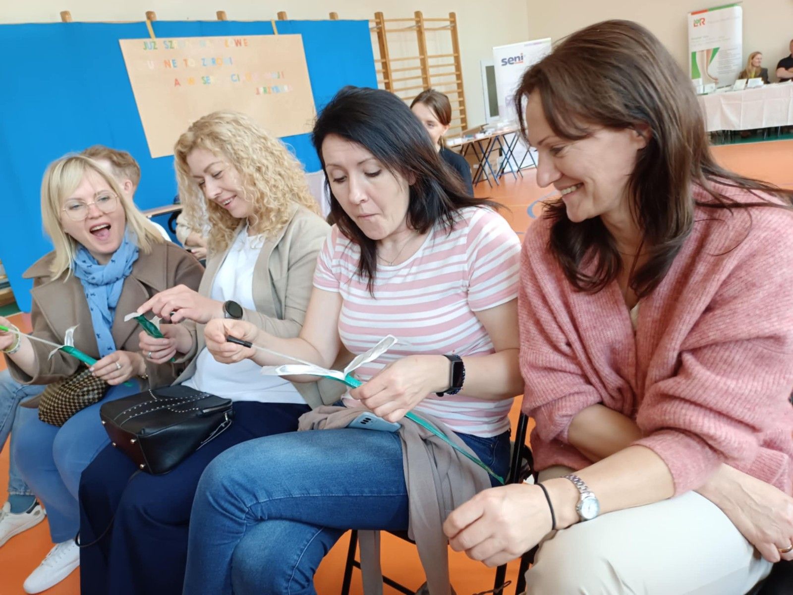 Uczestnicy warsztatów siedzący na hali sportowej z akcesoriami warsztatowymi.
