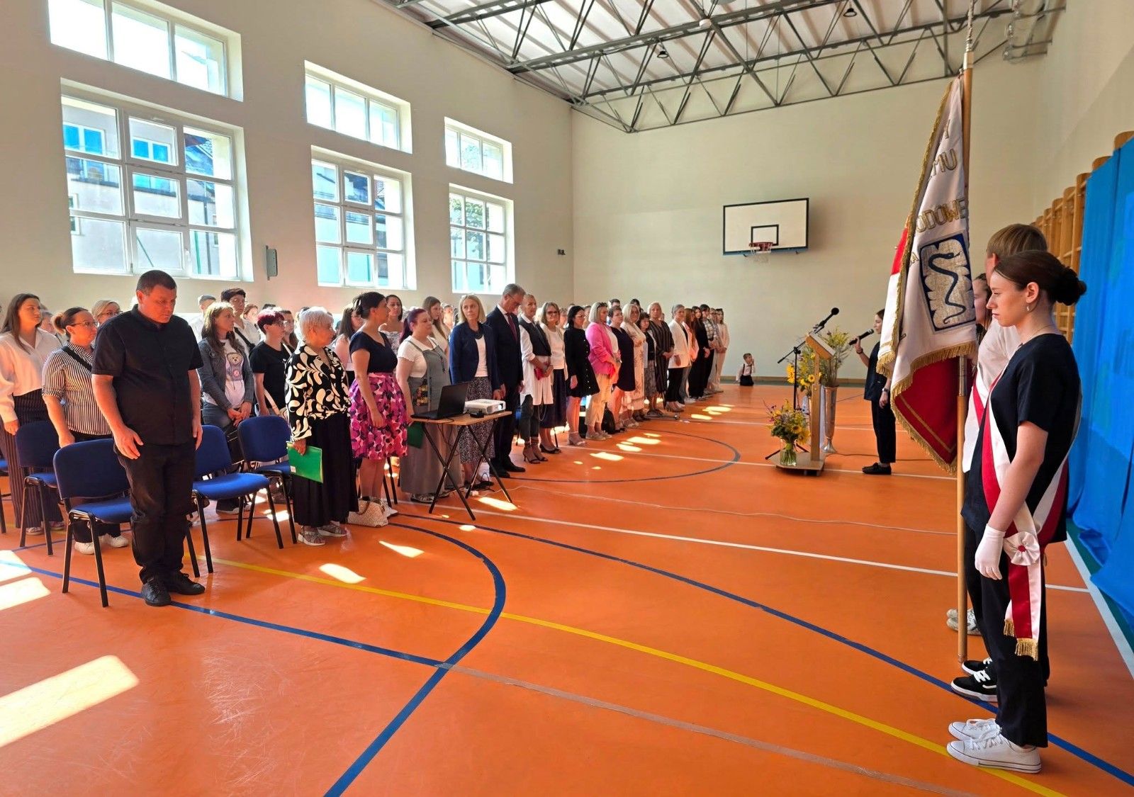 Widok na uczniów i nauczycieli na hali sportowej podczas przemówienia.